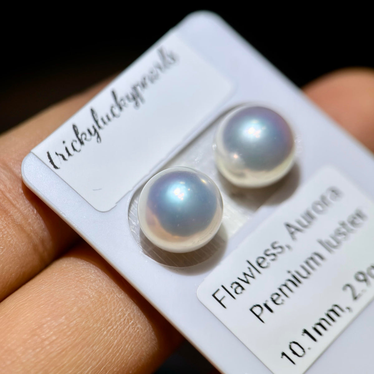 Two round lustrous pearl stud earrings mounted on a white display card held in a hand. The card has labels reading 'trickyluckypearls' and 'Flawless, Aurora Premium luster 10.