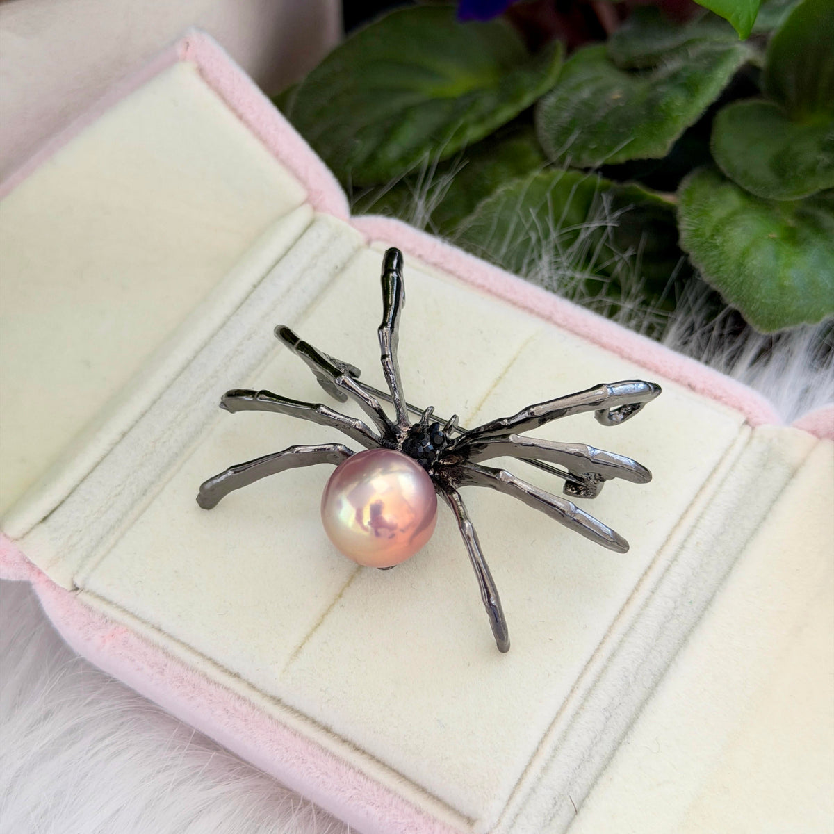 A spider-shaped brooch with metallic dark gray legs and a pink pearl center, displayed on an open light pink jewelry box with green leaves in the background.