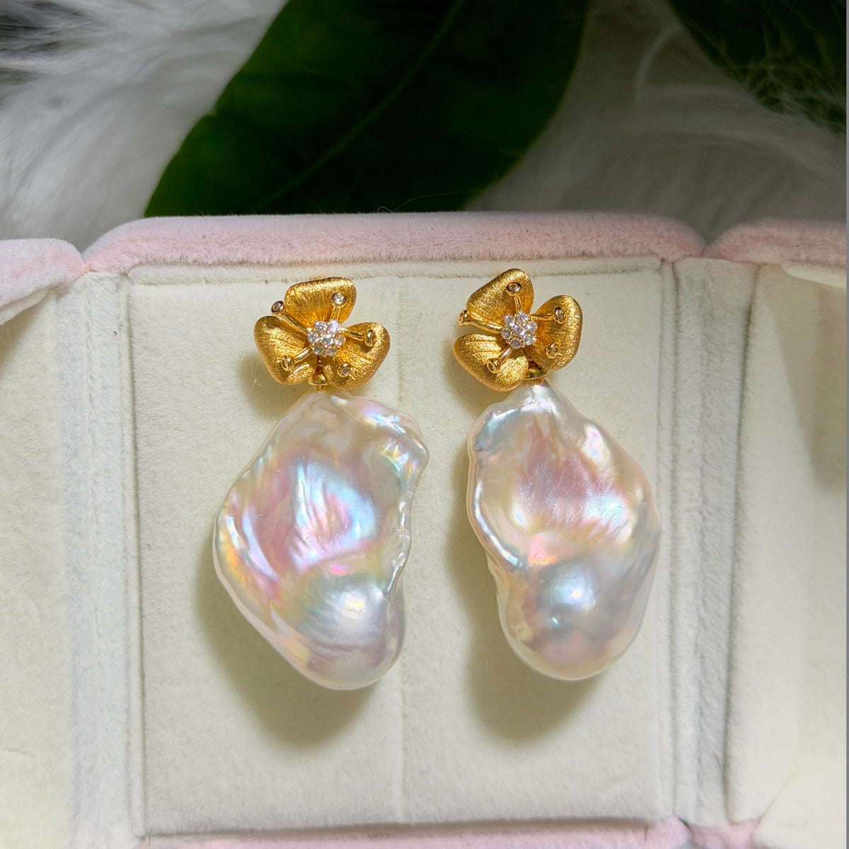 A pair of gold flower-shaped stud earrings with baroque pearls, displayed inside a light pink jewelry box.