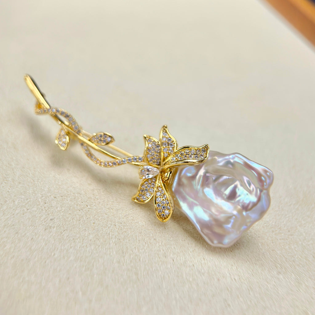 A gold rose-shaped brooch featuring a large iridescent pearl rosebud and a gold flower with pavé-set clear stones, displayed on a textured beige surface.