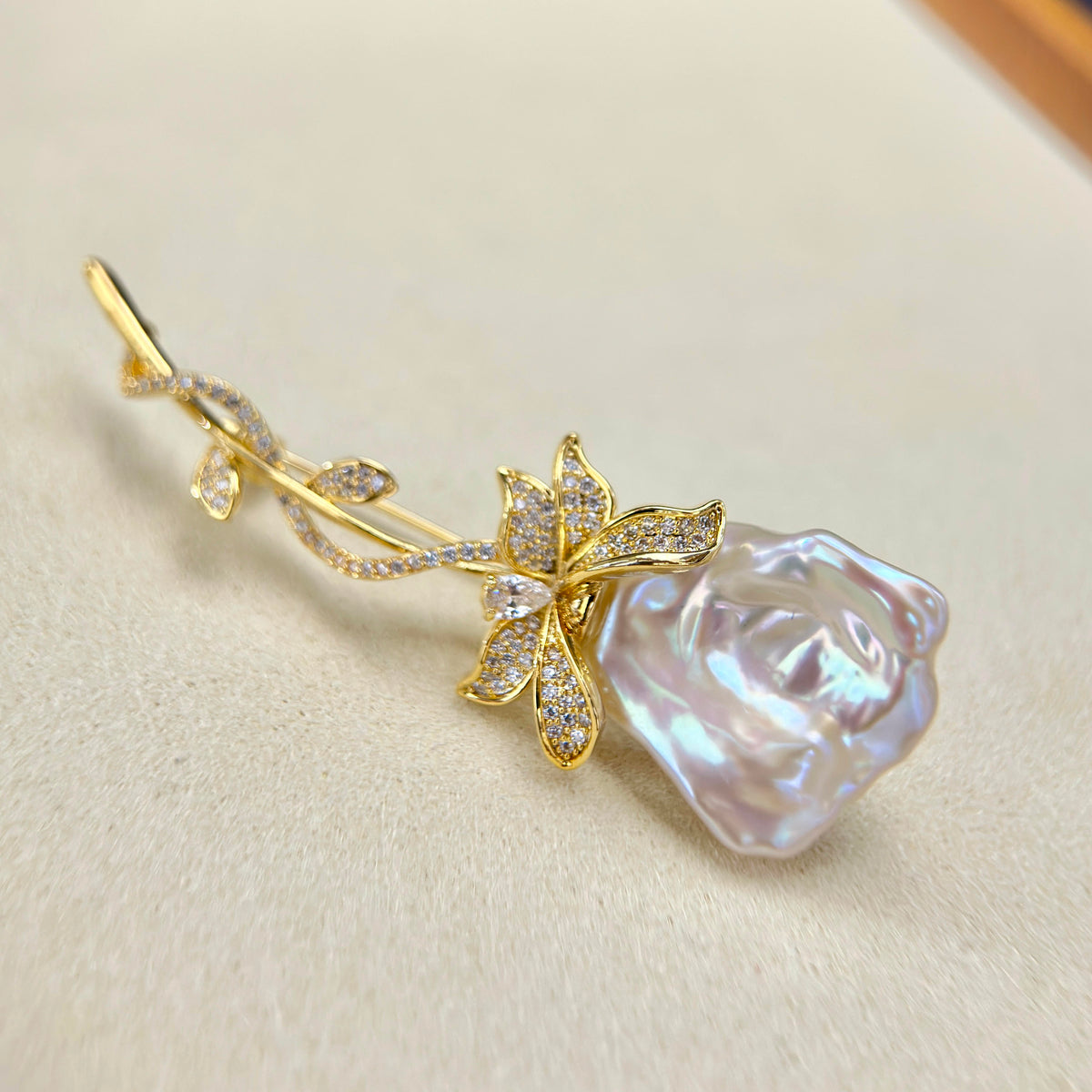 A gold rose-shaped brooch featuring a large iridescent pearl rosebud and a gold flower with pavé-set clear stones, displayed on a textured beige surface.