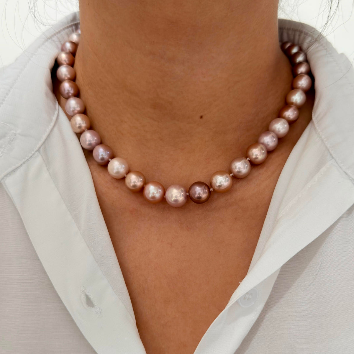 A close-up of a person wearing a strand of irregular baroque pearls in a dusty rose and lavender color, styled over a white collared shirt.