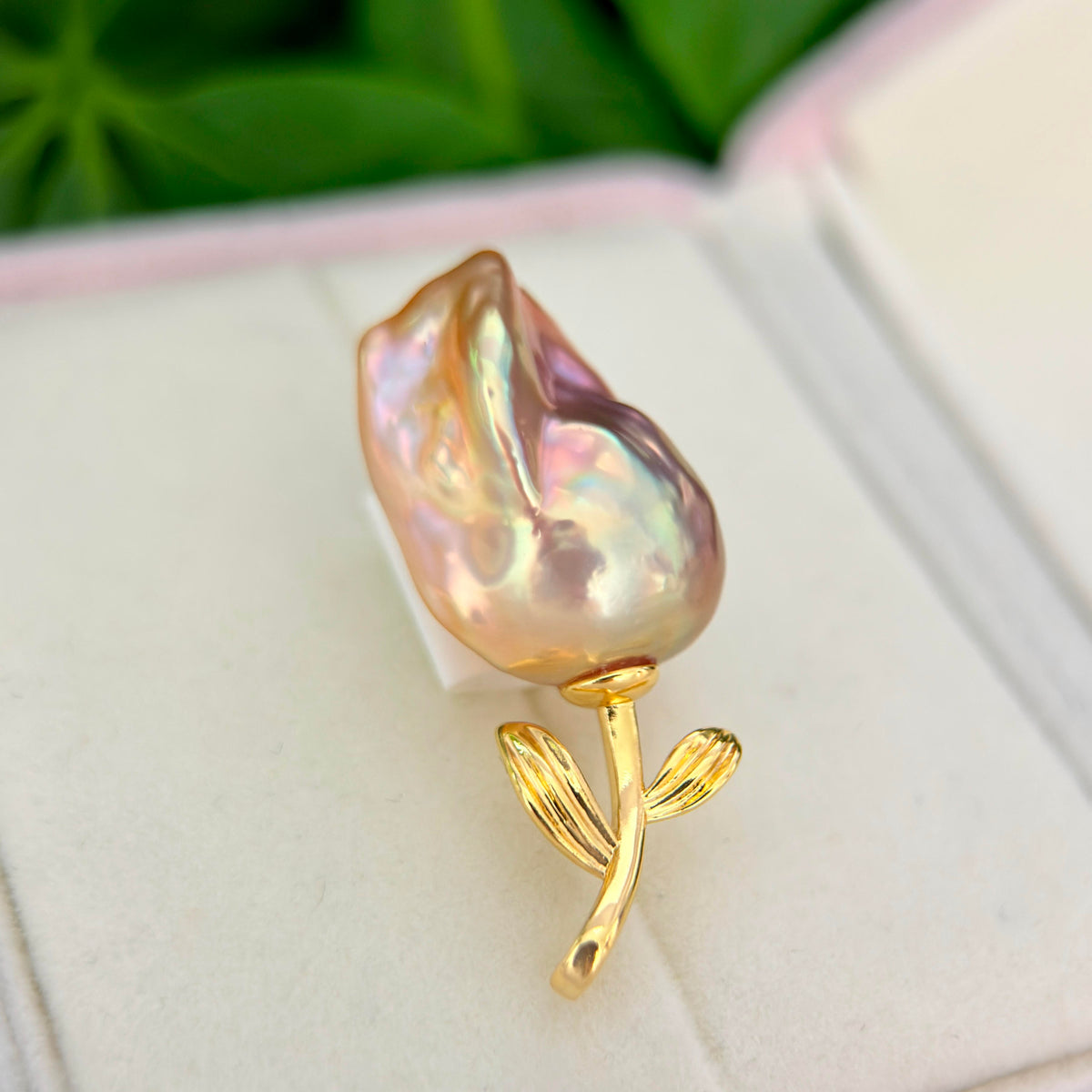 A close-up of a flower-shaped pearl brooch with a gold stem and leaves, resting on a white jewelry box.