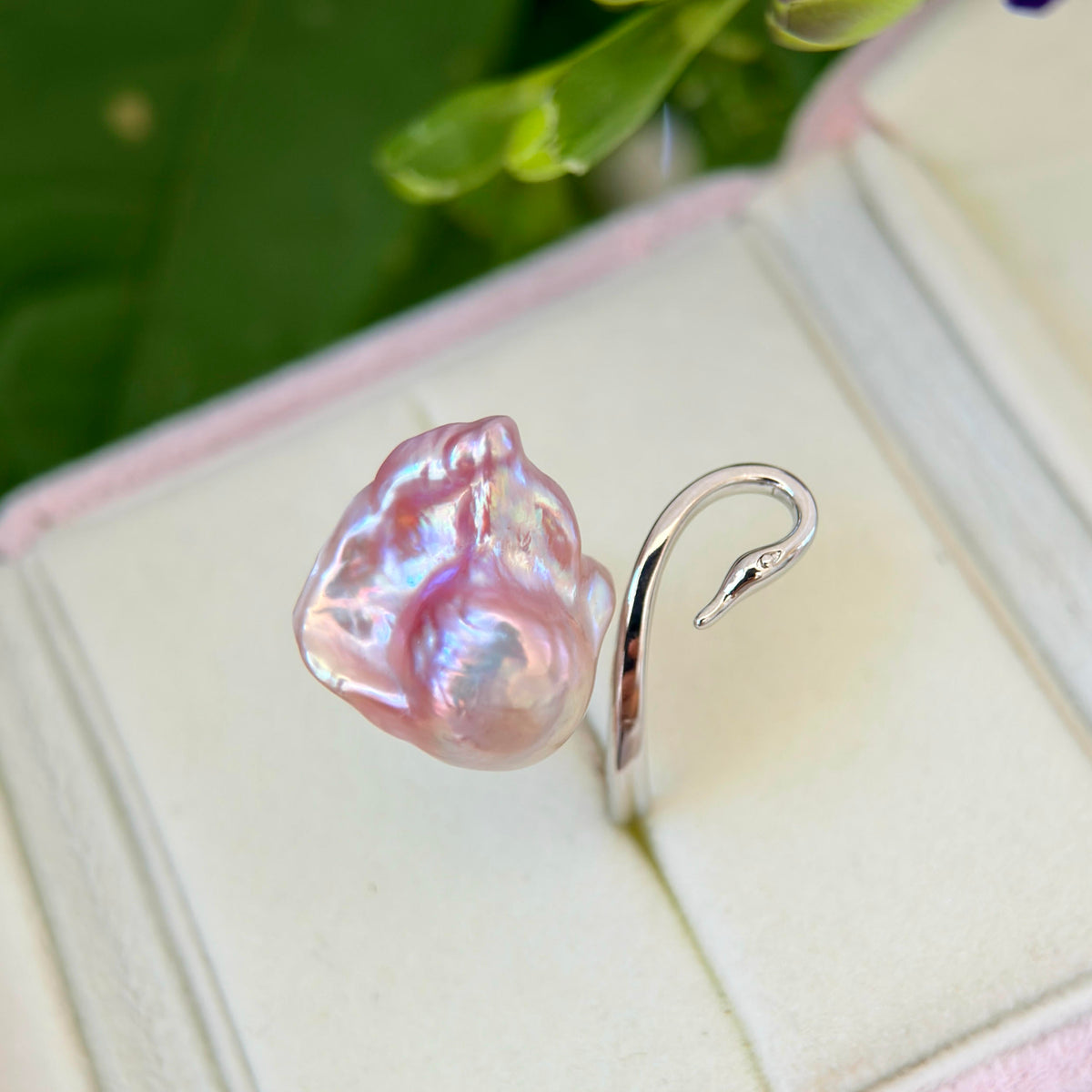 A silver ring featuring a large, irregular baroque pearl with iridescent pink and lavender hues, displayed on a light cream jewelry box insert with blurred green leaves in the background.
