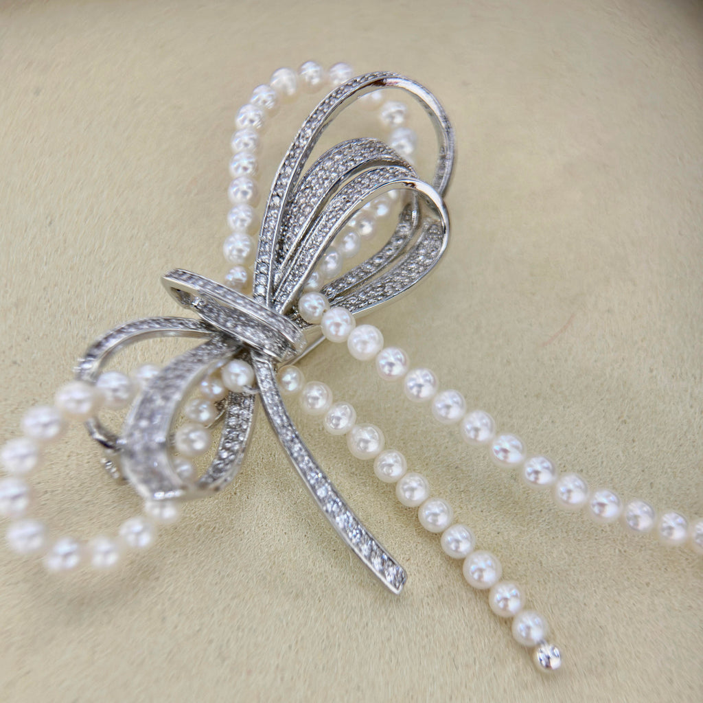 A silver-toned bow-shaped brooch embellished with clear rhinestones and draped with strands of white faux pearls, displayed on a textured beige background.