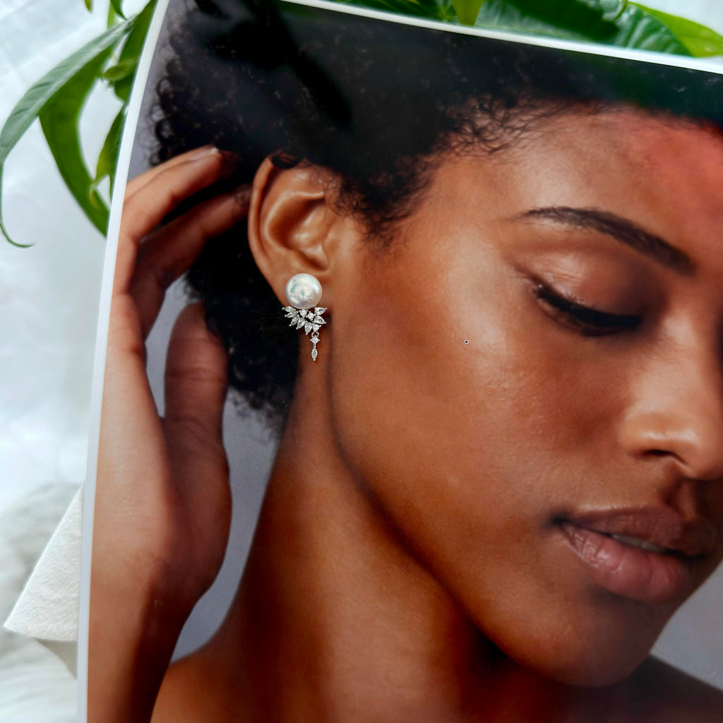 A close-up of a model wearing a silver-toned dangle earring with a white pearl and clear rhinestone accents, shown against a soft white background with green leaves at the top.