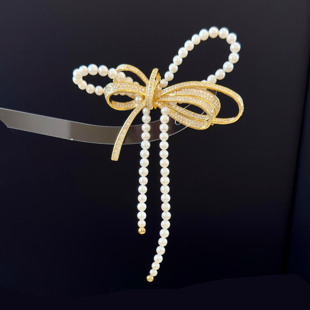 A decorative bow-shaped hair accessory featuring white faux pearls and a gold-toned center with small clear stones, displayed against a dark background.