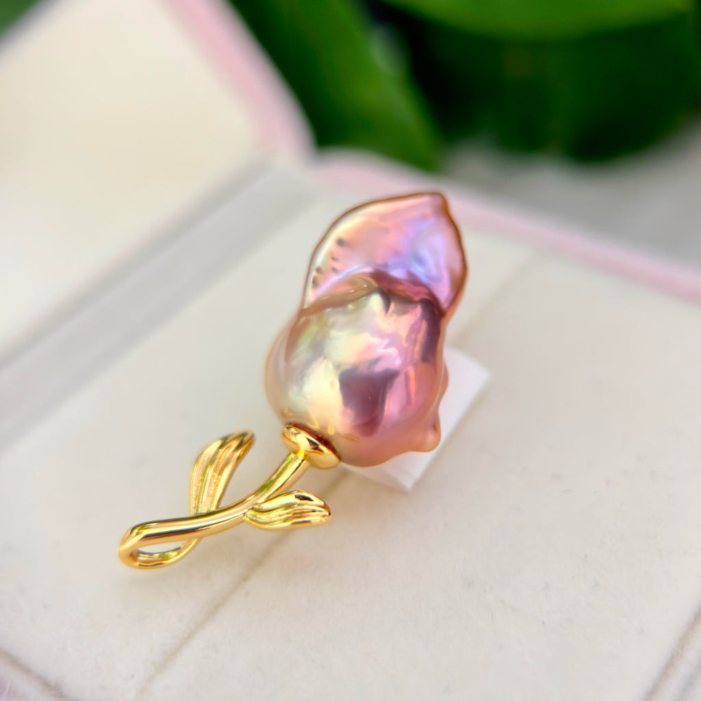 A close-up of a gold flower-shaped brooch featuring a baroque pearl with iridescent pink, purple, and green hues, displayed on a white jewelry box.