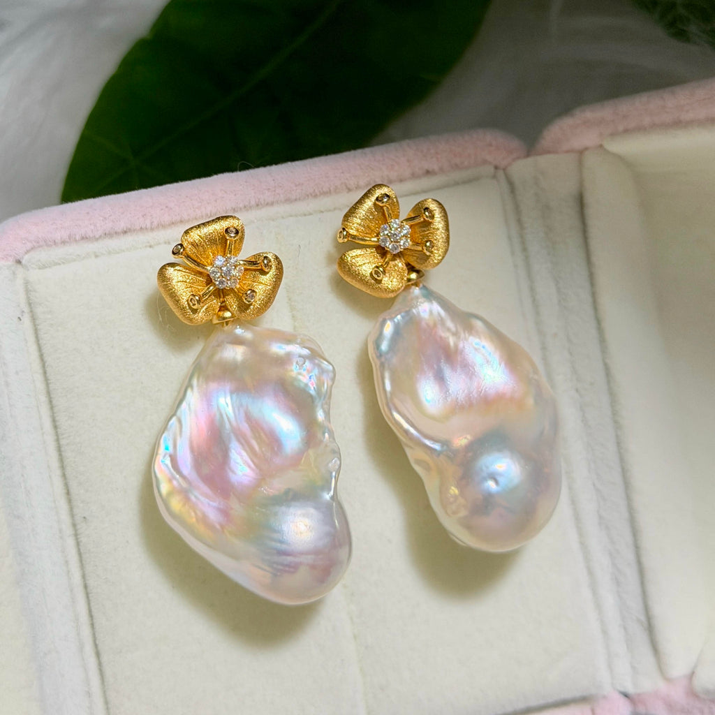 A pair of gold flower-shaped stud earrings with small clear stones and large, irregular iridescent pearl drops, displayed on a pink-lined jewelry box.