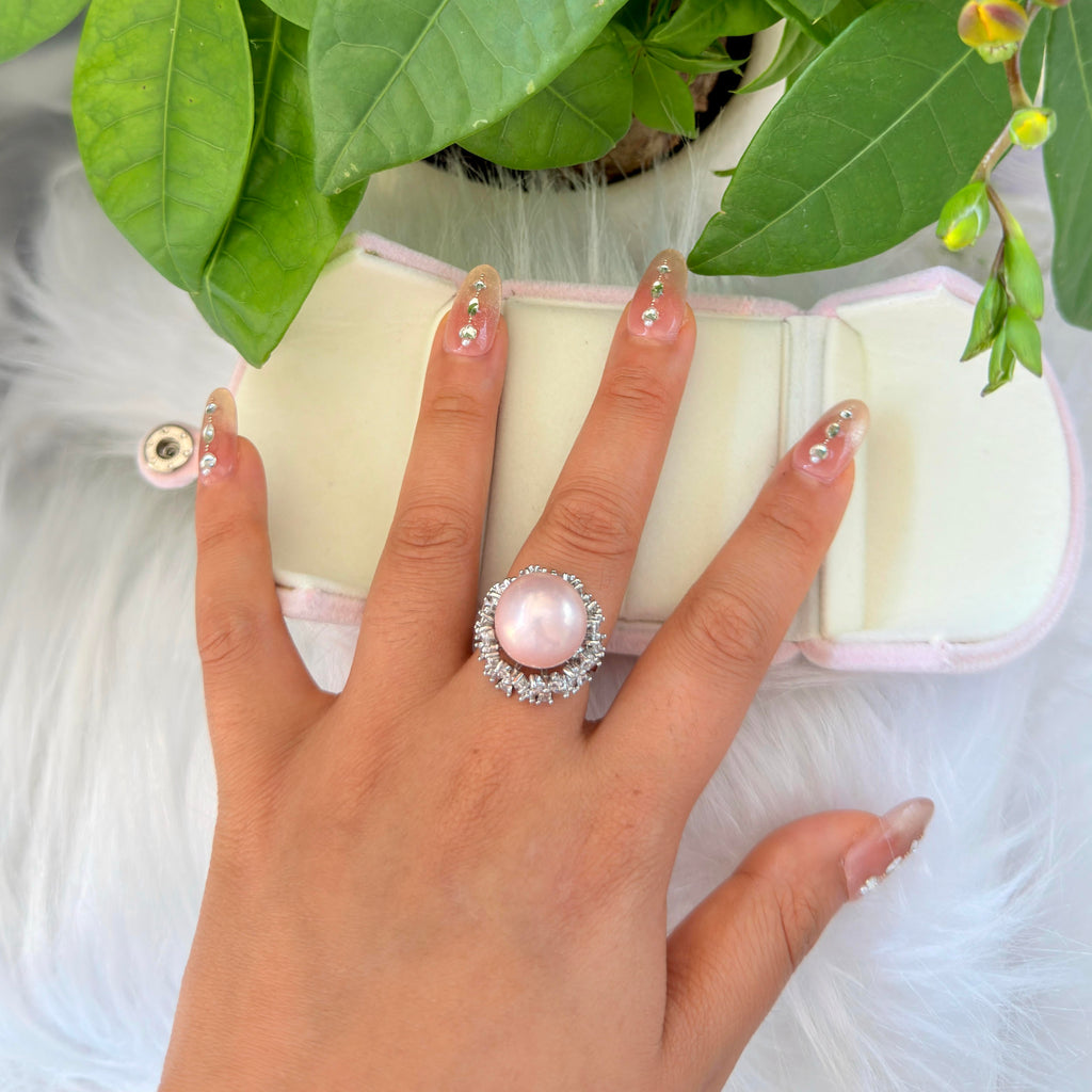 A hand with decorated nails wears a silver ring featuring a large pink pearl surrounded by small clear stones, resting on a white faux fur surface next to an open cream jewelry box and green leaves.