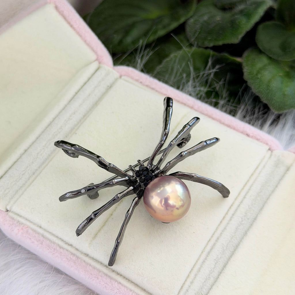 A spider-shaped brooch with a glossy iridescent pearl and dark metallic legs, displayed on an open pink-lined jewelry box with green foliage in the background.