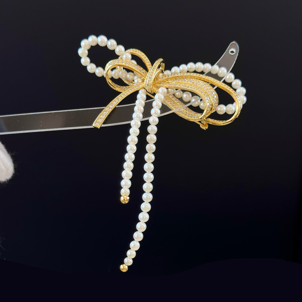 A decorative bow-shaped hair accessory with white pearl strands and a gold-toned metal bow, shown against a dark background.