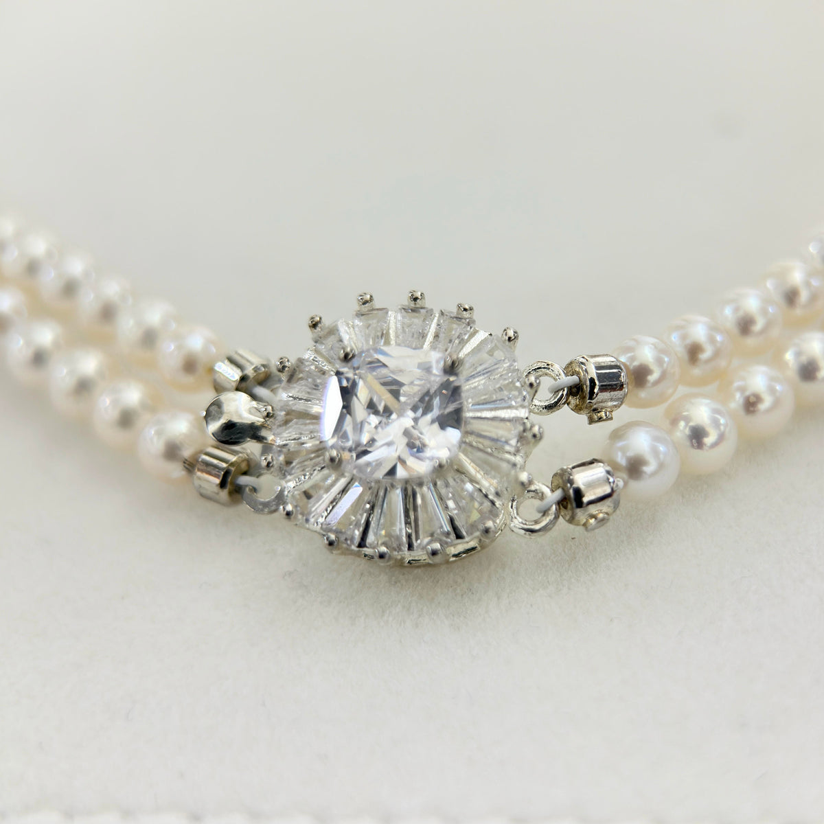 A close-up of a pearl necklace with a central clear gemstone cluster in a silver-toned setting, photographed on a soft light background.
