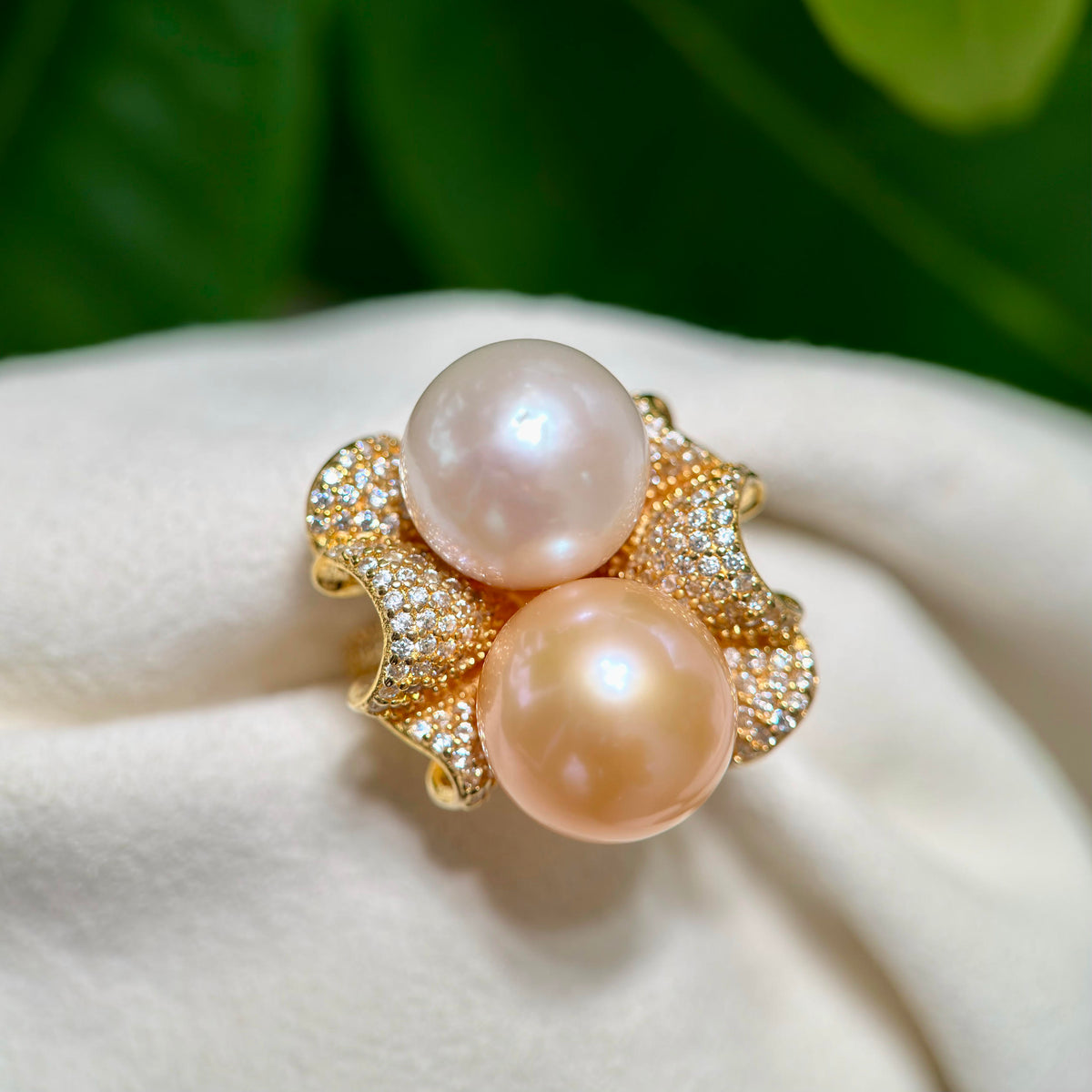 A gold ring featuring two large, round pinkish pearl-like beads set in a floral, pavé-set band, resting on a soft white fabric surface with a blurred green background.
