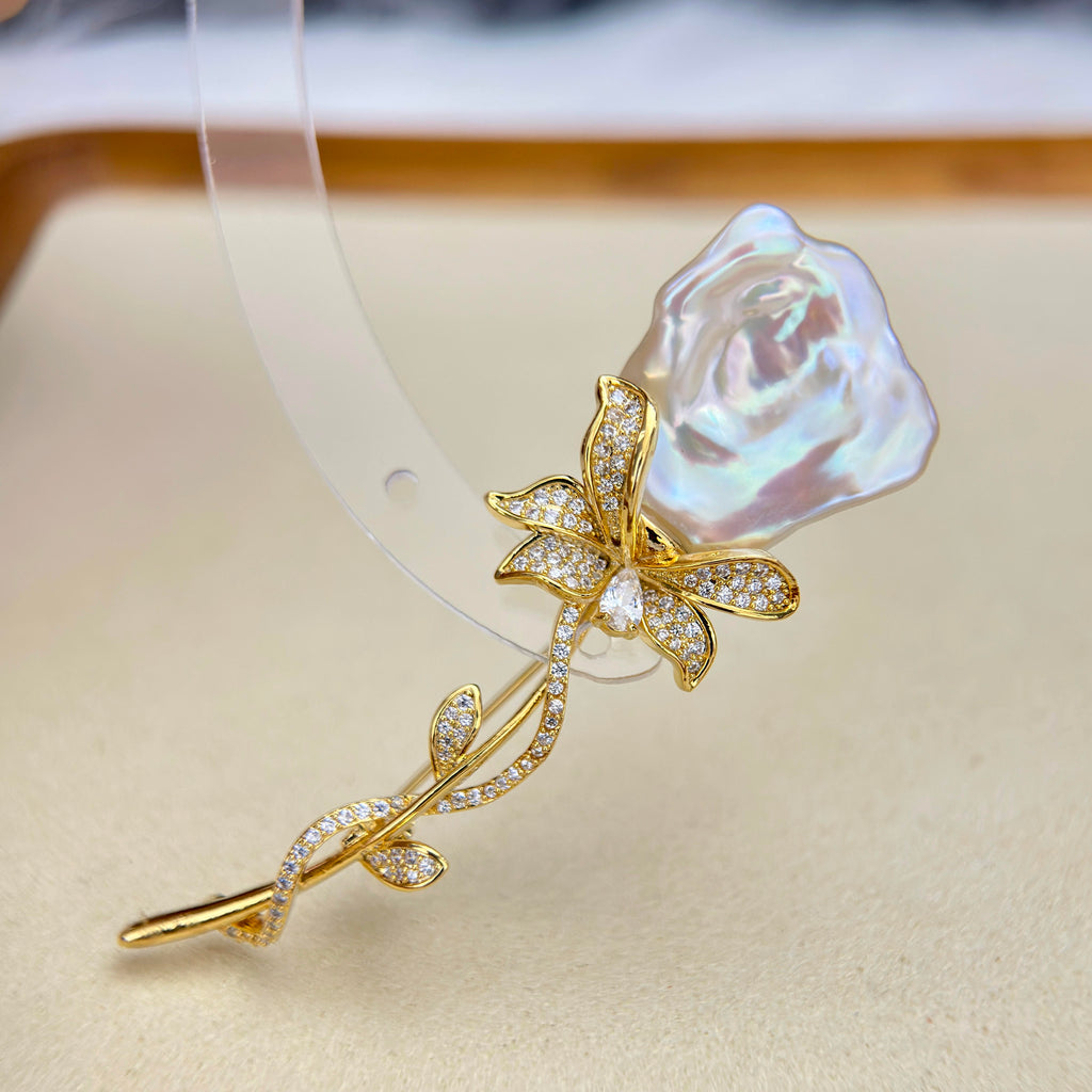 A gold-tone rose-shaped hair claw clip featuring a large, iridescent pearl-like rosebud and a stem paved with small clear stones, displayed on a beige surface.