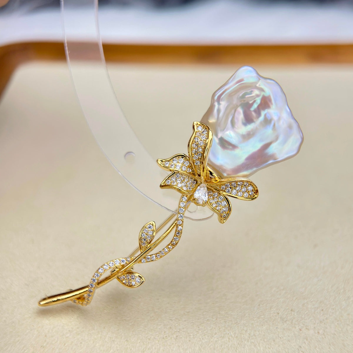 A gold-tone rose-shaped hair claw clip featuring a large, iridescent pearl-like rosebud and a stem paved with small clear stones, displayed on a beige surface.