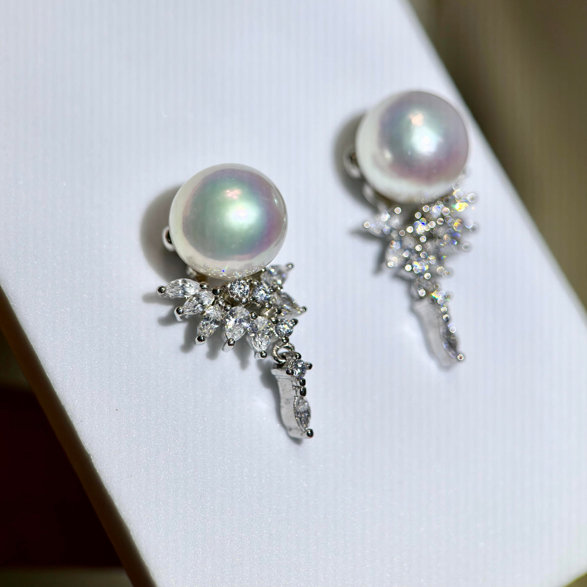 A pair of pearl and crystal drop earrings displayed on a white jewelry stand. Each earring features a round iridescent white pearl set in silver-toned metal, with a cluster of clear marquise and round crystals extending downward.