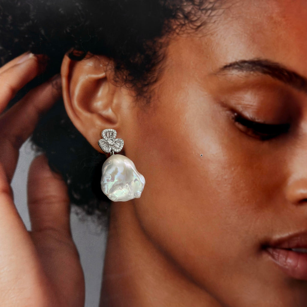 A close-up of a model wearing a silver-toned dangle earring with a small floral cluster of clear stones and a large, irregular white baroque pearl drop.
