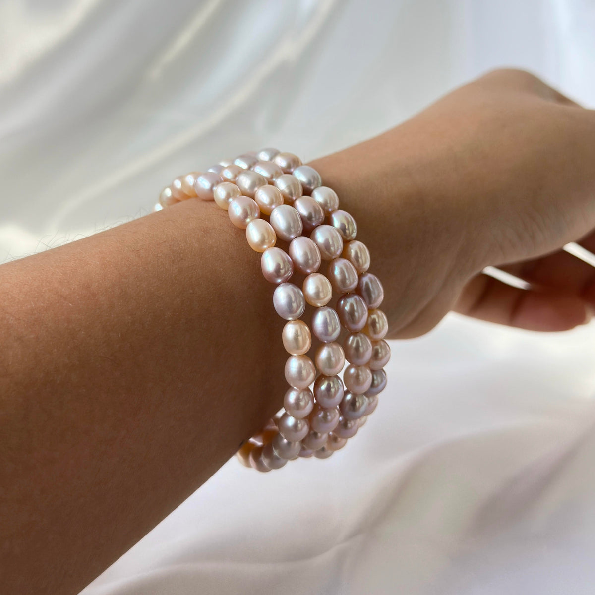 A close-up of a person's wrist wearing a multi-strand bracelet made of irregular, lustrous freshwater pearls in soft pink, cream, and white tones, set against a soft white draped fabric background.