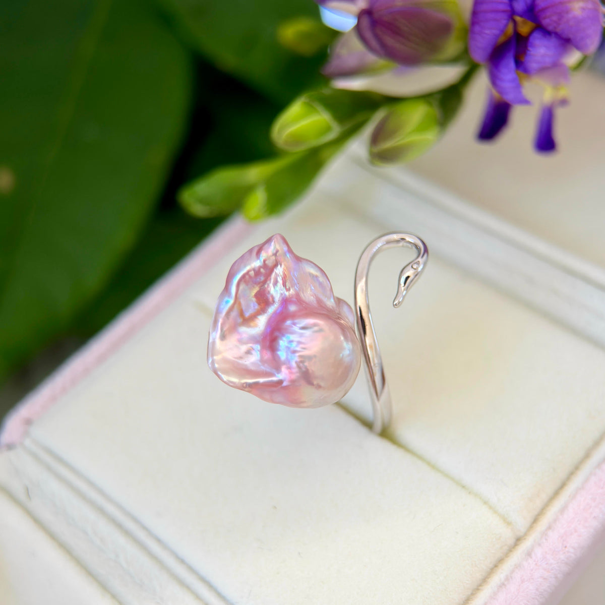 A silver ring with a large, irregularly shaped pink baroque pearl featuring iridescent blue and purple flashes, displayed on a light pink velvet ring box with green leaves and purple flowers in the background.