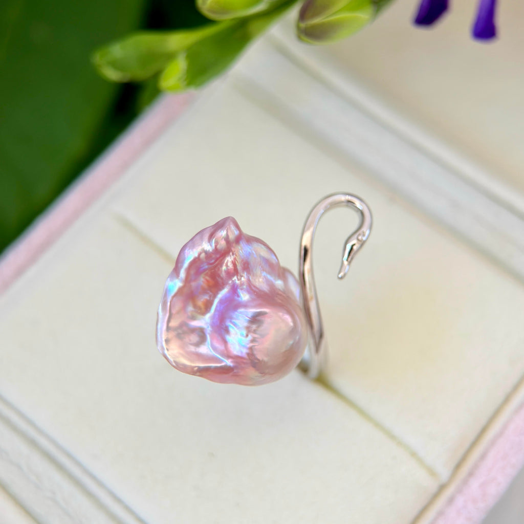 A silver ring featuring a large, baroque-style pink iridescent pearl, displayed on a cream-colored ring box with blurred green leaves in the background.