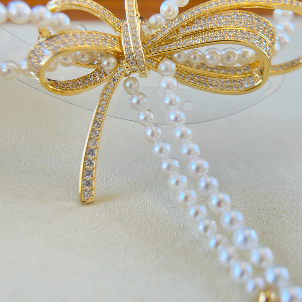 A close-up of a gold bow-shaped jewelry piece adorned with pavé-set clear stones and draped strands of white faux pearls on a light beige surface.