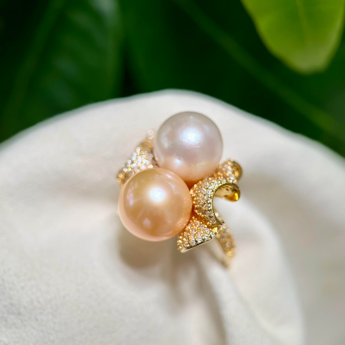 A gold ring featuring two large round freshwater pearls in soft peach and cream tones, accented with pavé-set clear stones, displayed on a white textured surface with blurred green foliage in the background.
