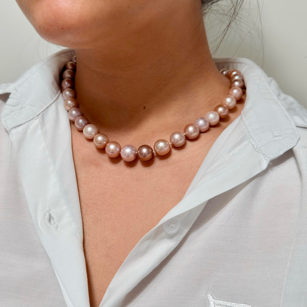A close-up of a person wearing a single strand of lustrous pinkish pearl necklace with irregular oval beads, styled with a white collared shirt.