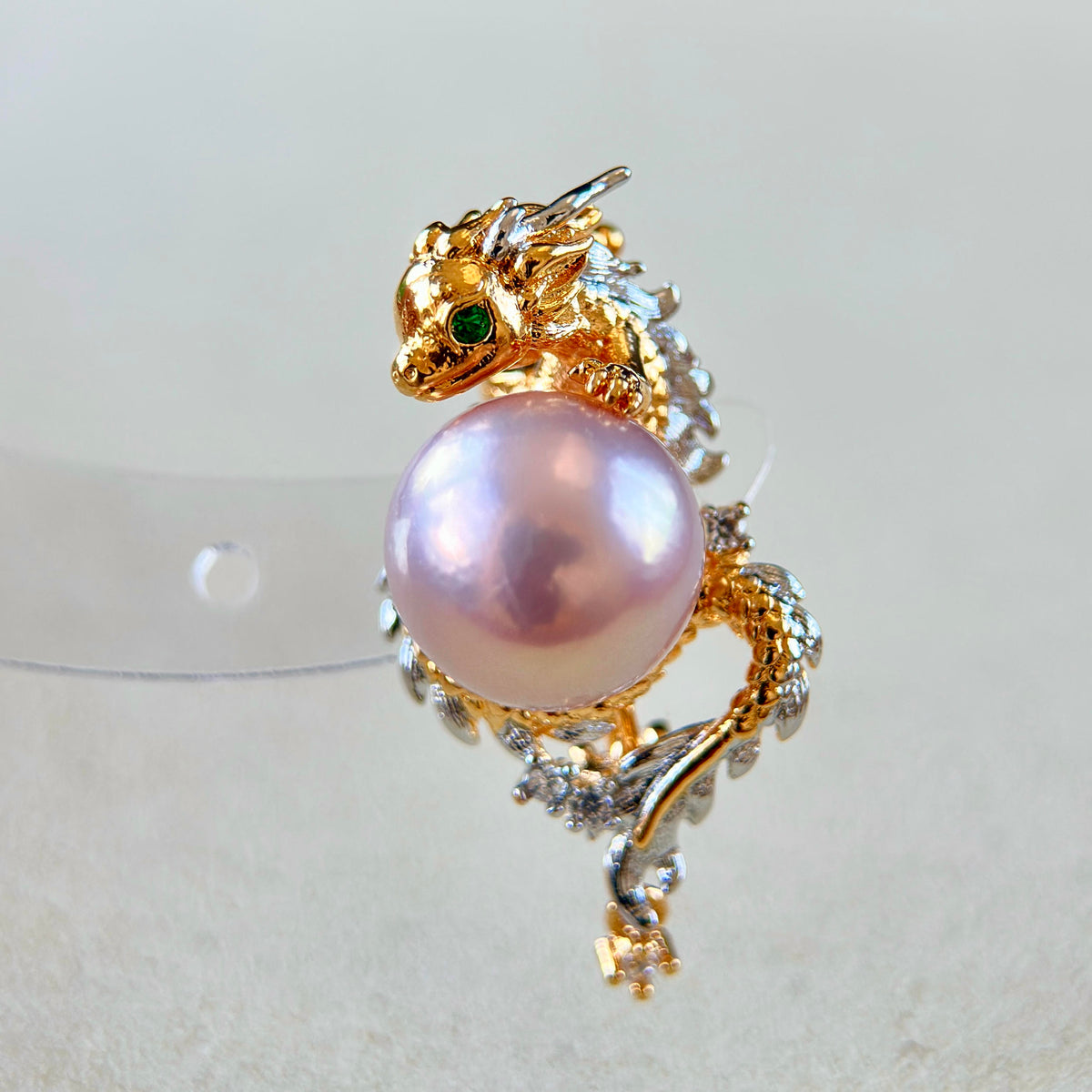 A gold dragon ring featuring a large pink pearl, small green gemstone eyes, and silver-toned crystal accents, displayed on a light beige surface.