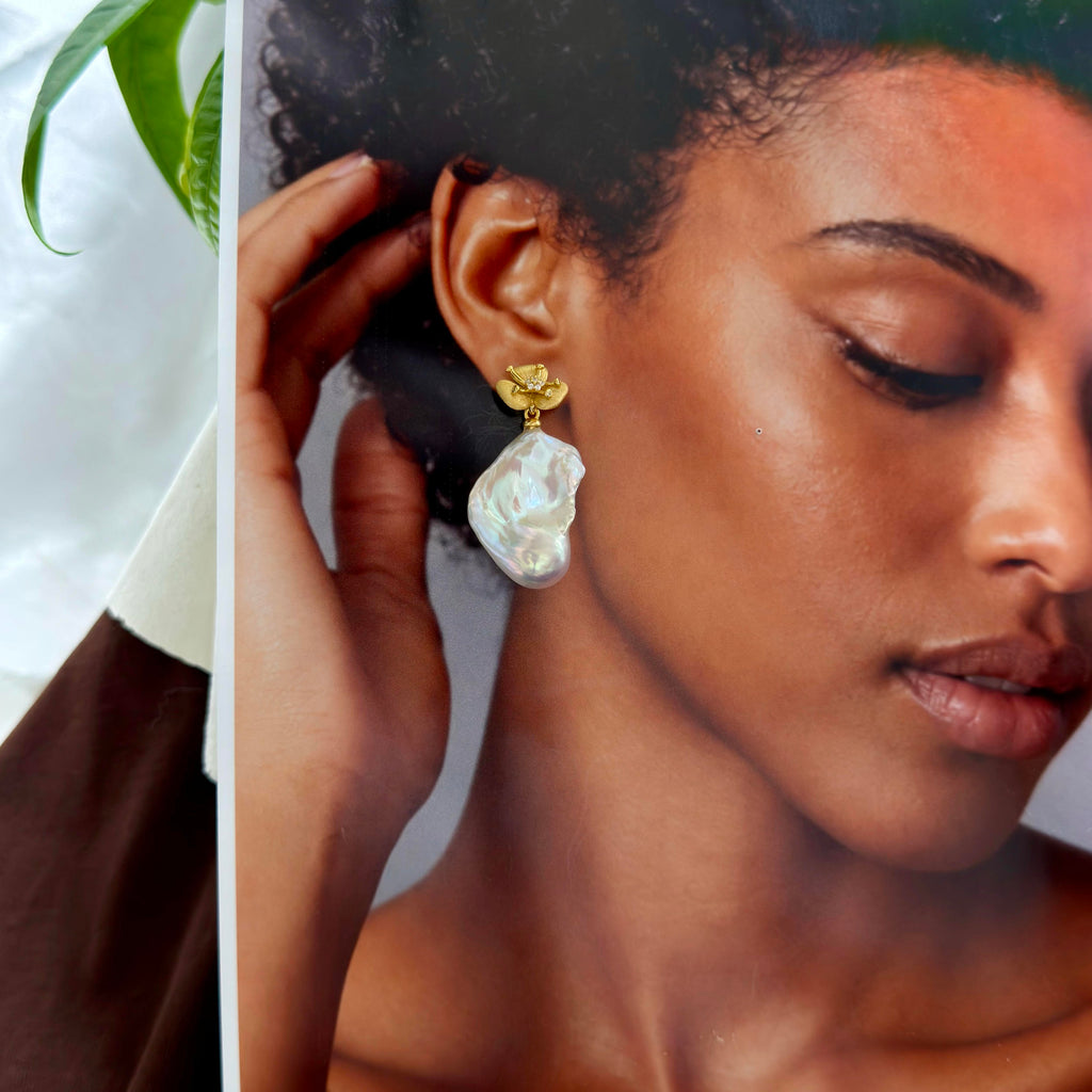 A close-up lifestyle shot of a model wearing a gold drop earring with an irregular, milky white pearl-like stone. The photo is cropped tightly, with the model's face and hand visible, and a sliver of a white background with green leaves on the left edge.
