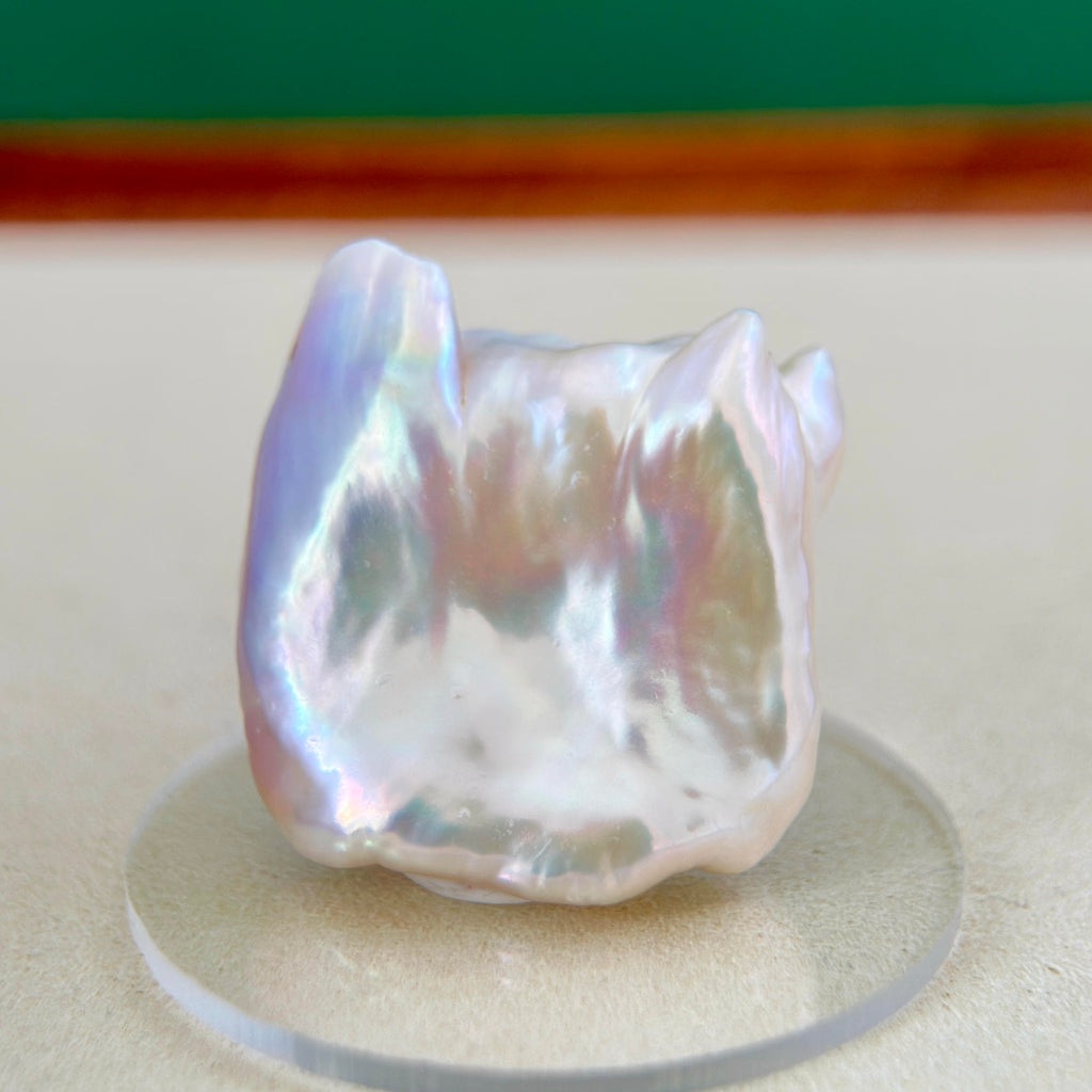 A baroque freshwater pearl with a pearlescent white body and iridescent rainbow sheen, displayed on a clear circular stand.