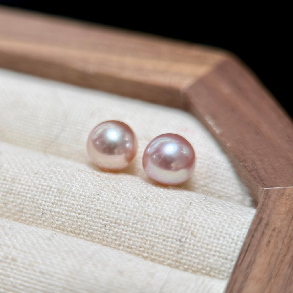 Two round pink freshwater pearls displayed on a cream fabric-lined jewelry box with a wooden frame.