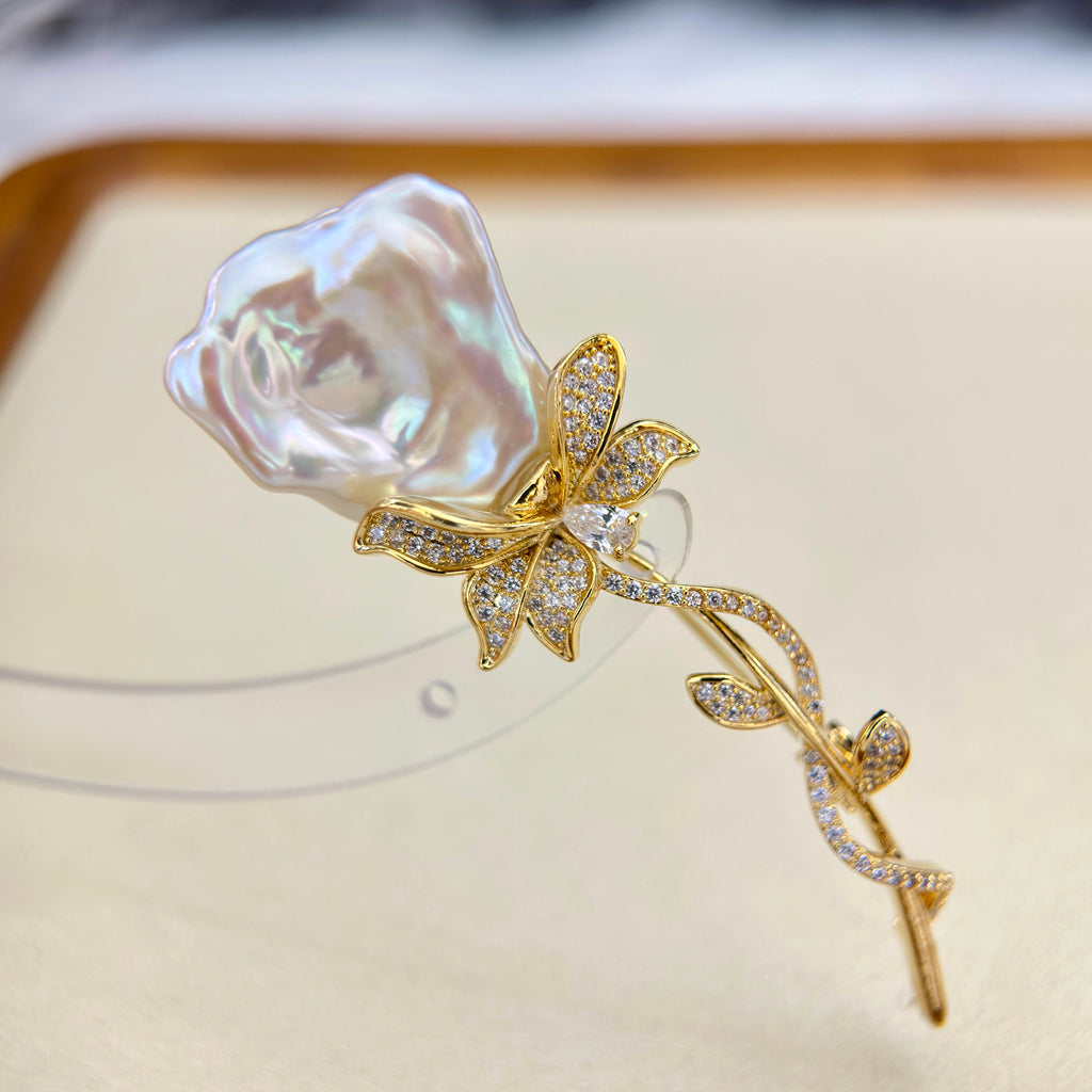 A gold rose-shaped brooch featuring a large, irregular lustrous white pearl flower head and pavé-set clear stones on the stem and leaves, displayed on a beige jewelry stand.