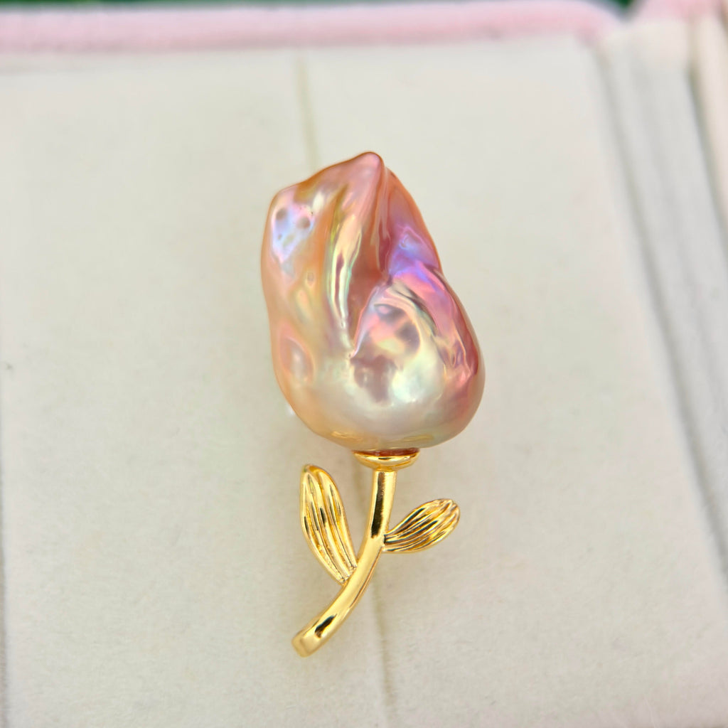 A baroque pearl rose brooch with a pearlescent pink, purple, and gold iridescent flower head and a polished gold stem, displayed on a light cream jewelry box.