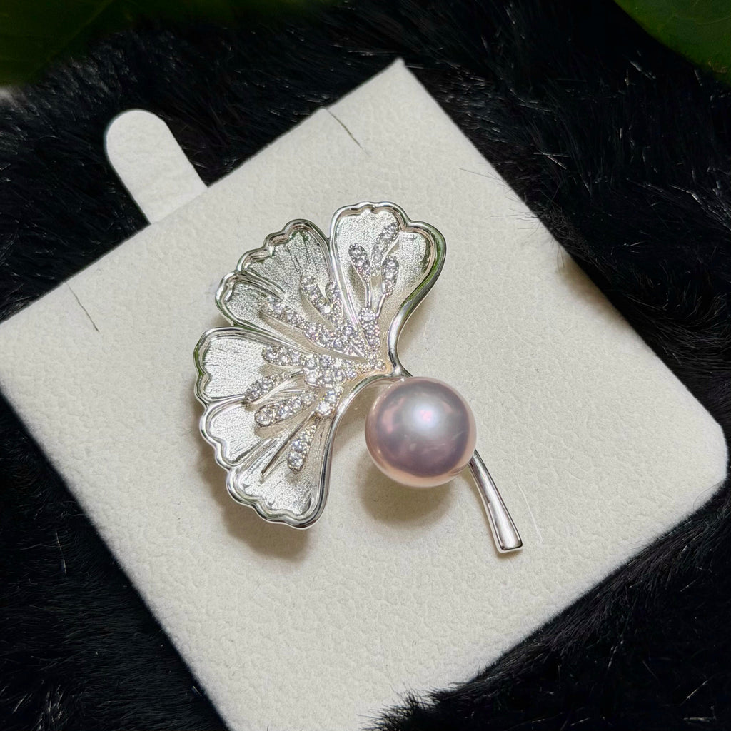 A silver ginkgo leaf brooch with a lustrous pink pearl and small clear stones, displayed on a white jewelry card against a dark textured background.