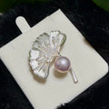 A silver ginkgo leaf brooch with a lustrous pink pearl and small clear stones, displayed on a white jewelry card against a dark textured background.
