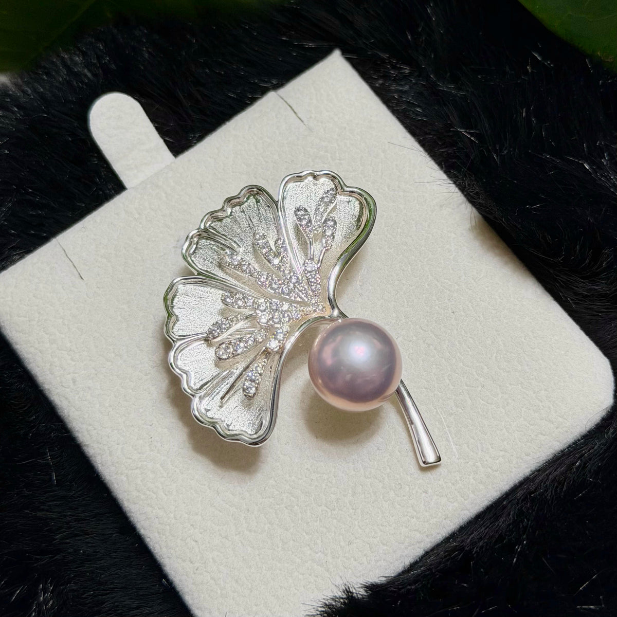 A silver ginkgo leaf brooch with a lustrous pink pearl and small clear stones, displayed on a white jewelry card against a dark textured background.