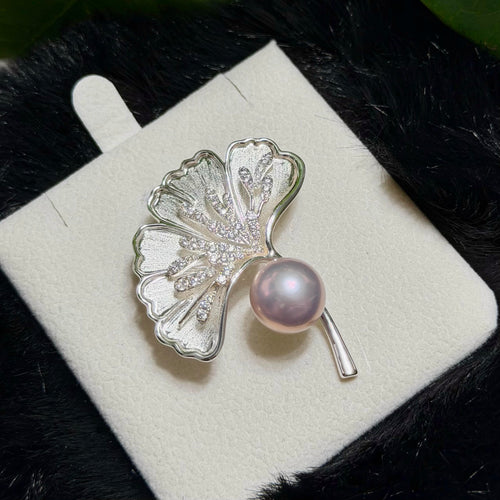 A silver ginkgo leaf brooch with a lustrous pink pearl and small clear stones, displayed on a white jewelry card against a dark textured background.