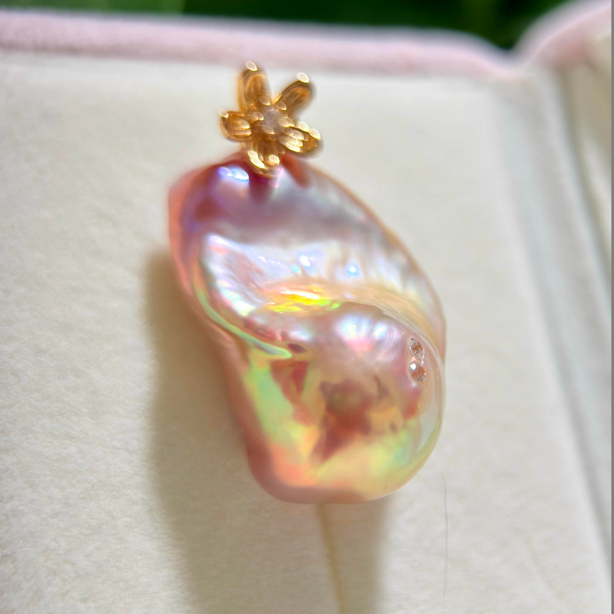 A baroque pearl pendant with an iridescent pink and green sheen, featuring a small gold flower-shaped bail, displayed on a cream-colored jewelry box.