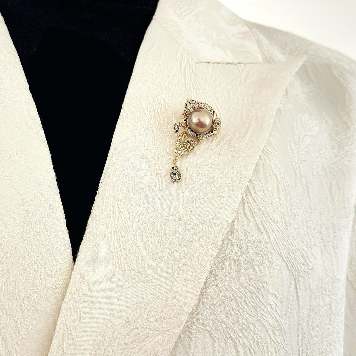 A close-up of a textured cream-colored jacket with a decorative brooch pinned to the lapel. The brooch features a central pinkish pearl surrounded by gold-toned filigree and small clear stones, with a teardrop-shaped dangling element.