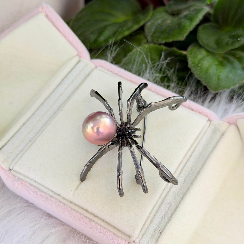 A spider-shaped brooch with a pink pearl bead and dark metallic legs, displayed on an open pale pink jewelry box with green leaves in the background.
