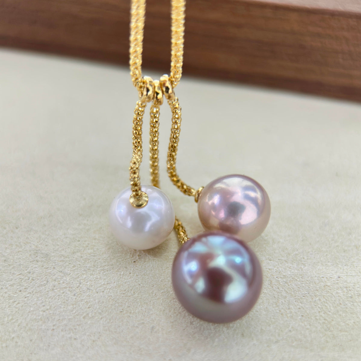 A close-up of a gold chain necklace with three dangling pearl-like beads in white, pale pink, and iridescent mauve tones, resting on a light beige surface.