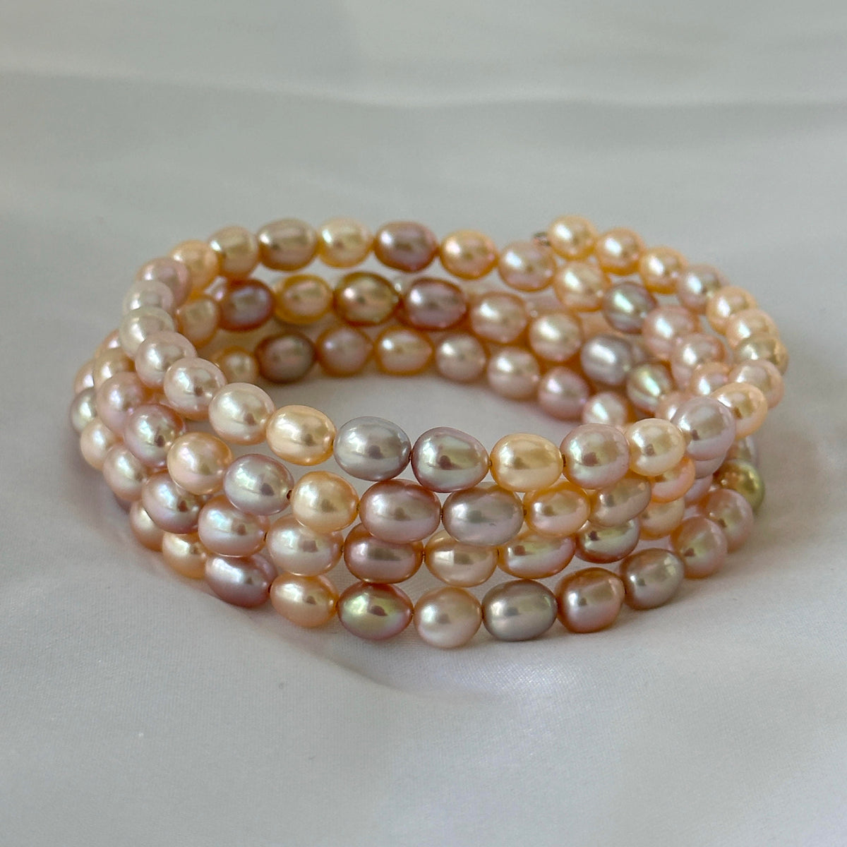 A multi-strand wrap bracelet made of irregular baroque pearls in soft peach, pink, champagne, and lavender tones, coiled on a light gray fabric background.