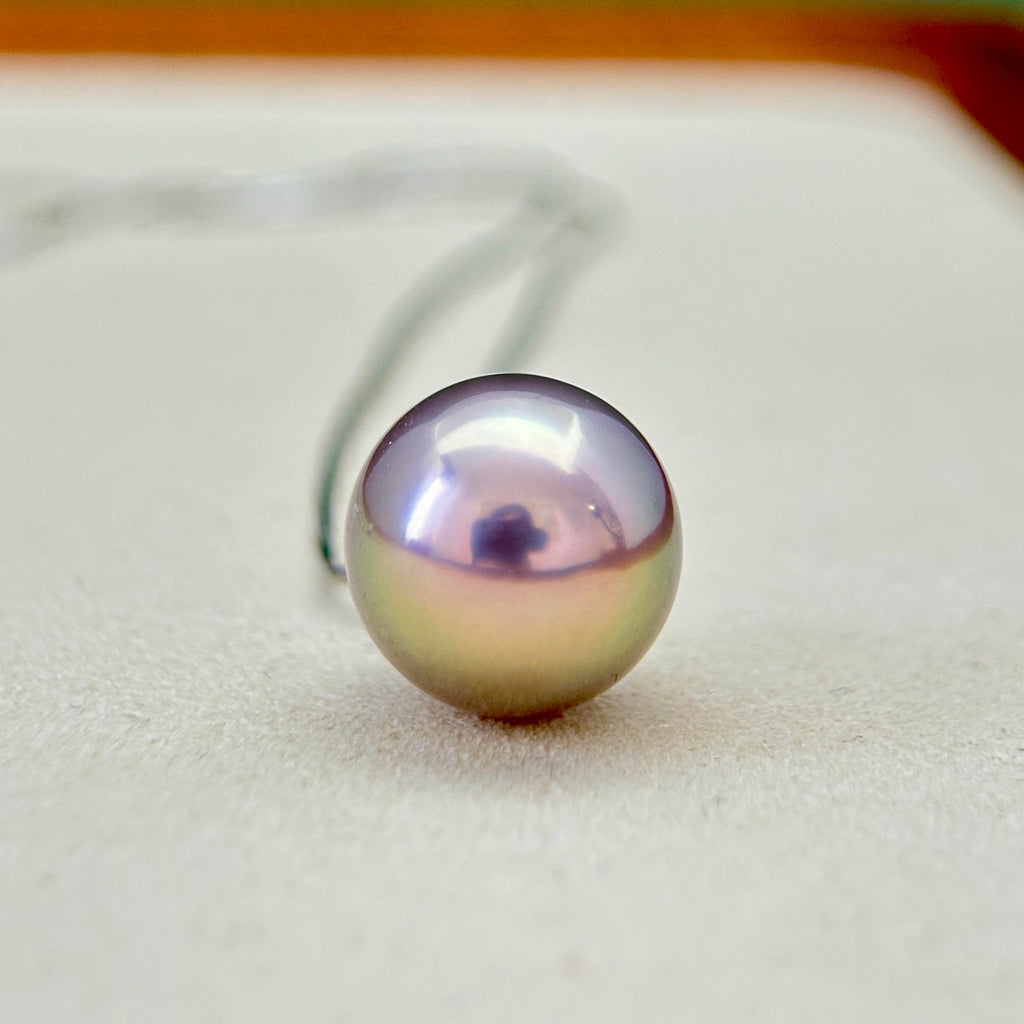 A single iridescent oval pearl pendant on a delicate silver chain, resting on a textured beige surface.
