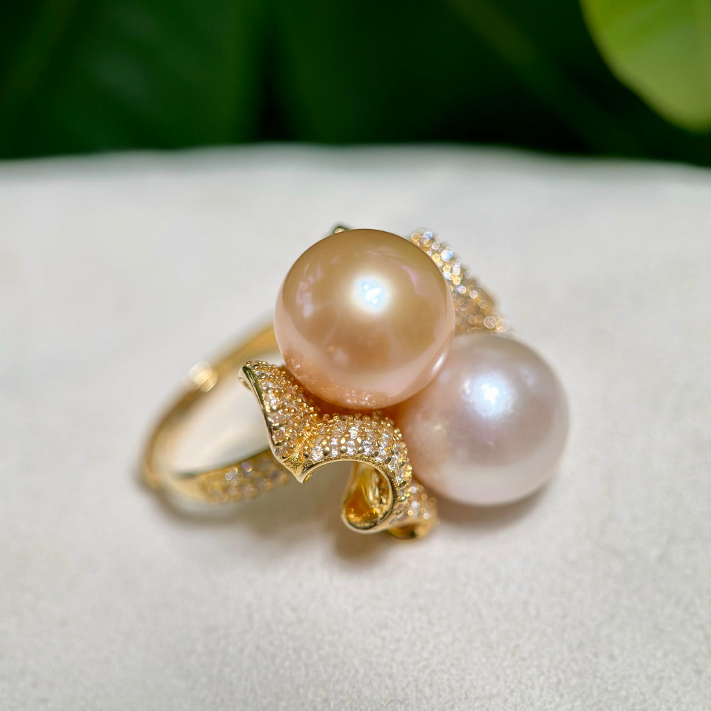 A gold ring featuring two large round pearls, one peachy gold and one pale pink, set on a textured white surface with a blurred green background.