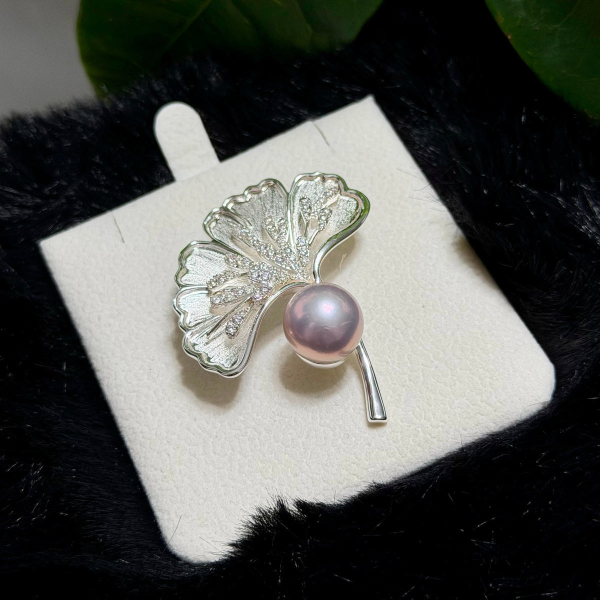 A silver ginkgo leaf brooch with a single lustrous pink pearl, displayed on a white jewelry card over a black textured surface with green leaves in the background.