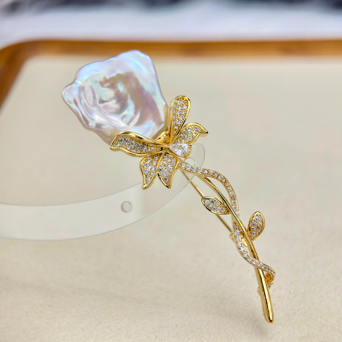 A gold flower-shaped hair claw clip decorated with pavé-set clear crystals and a large, lustrous white pearl rose at the top, displayed on a beige jewelry stand.