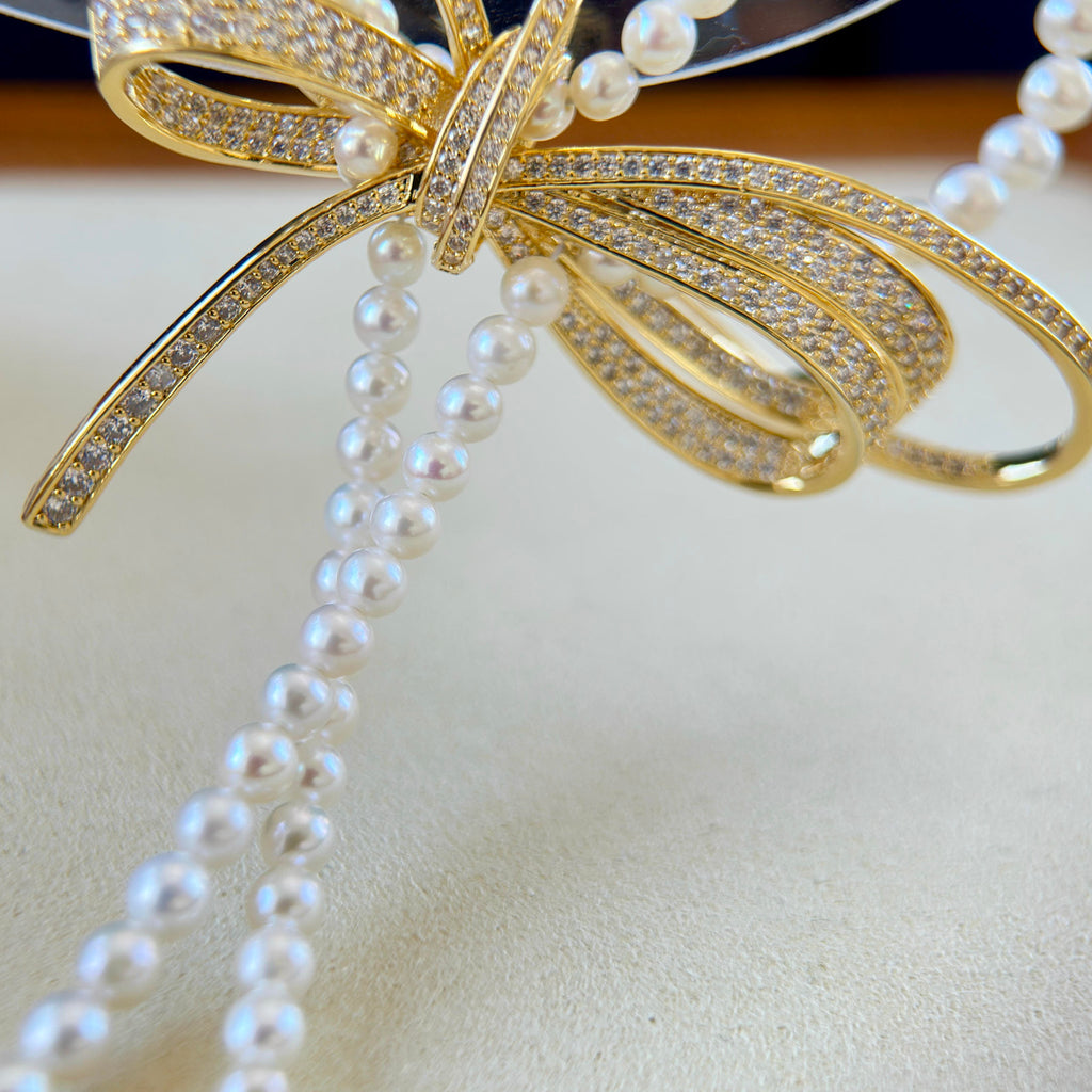 A close-up of a gold bow-shaped jewelry piece adorned with pavé stones and draped with strands of white faux pearls, resting on a light beige textured surface.
