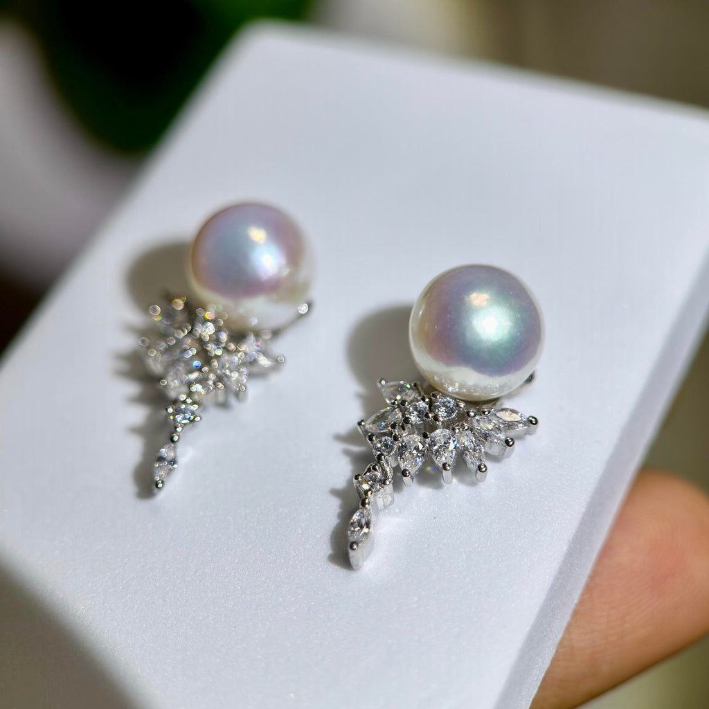 A pair of pearl and crystal drop earrings displayed on a white jewelry box, with a finger holding the box in the background.