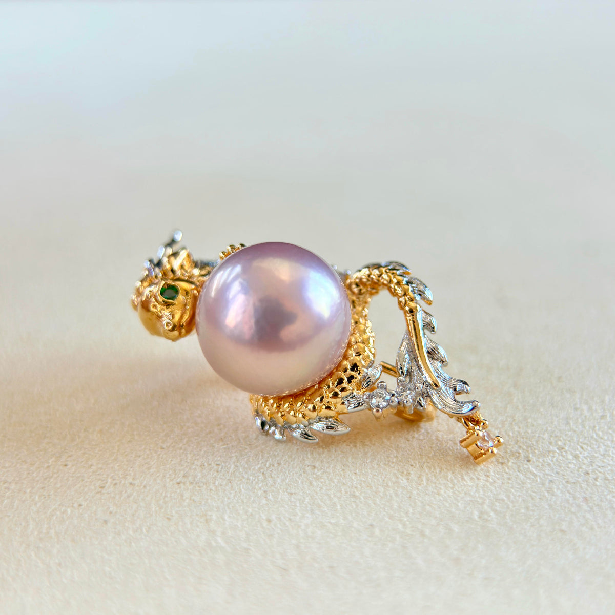 A gold ring designed as a stylized dragon wrapping around a large, lustrous pink pearl. The ring features small green emerald accents on the dragon's head and clear stones on its body, set against a soft beige background.